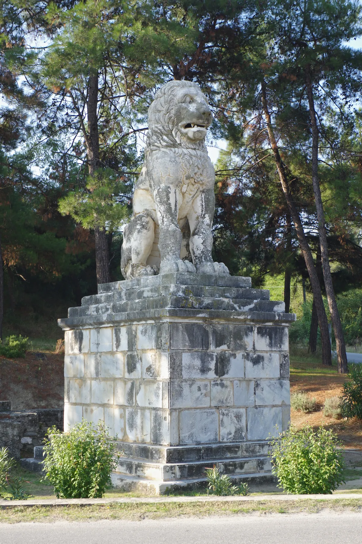 A large seated marble lion on a stone pedestal beside a road lined with trees